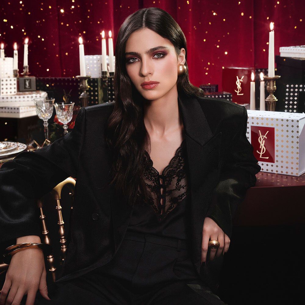 Model wearing a black suit and glam look, seated in a festive candlelit setting with YSL gift boxes.