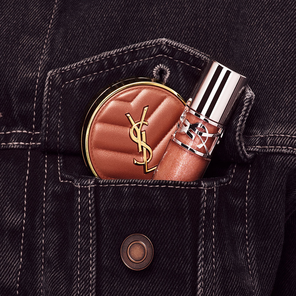 Hand placing YSL LOVESHINE Lip Oil Gloss and pink Make Me Blush compact into a denim pocket in shade Honey Moon.