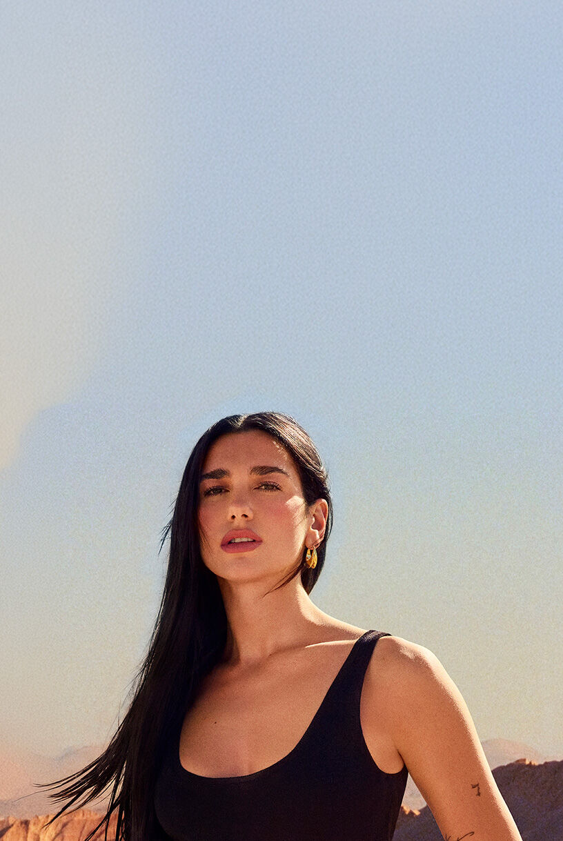 Dua Lipa, YSL Beauty ambassador, with flowing dark hair and a subtle tattoo, gazes directly at the camera in a desert setting.