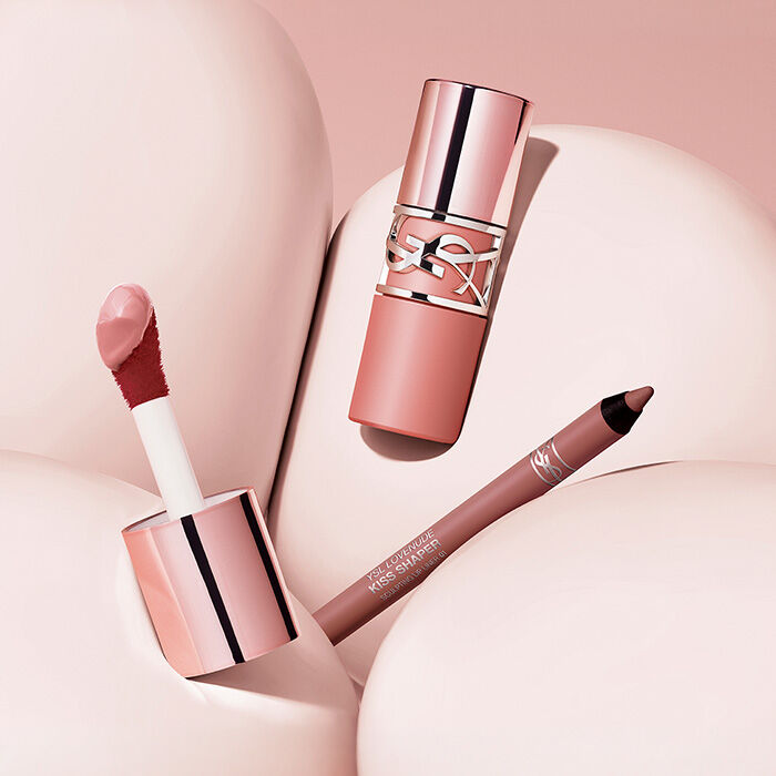 YSL LOVENUDE Kiss Shaper pencils and Lip Blusher lipsticks in various shades, arranged on a pink background.