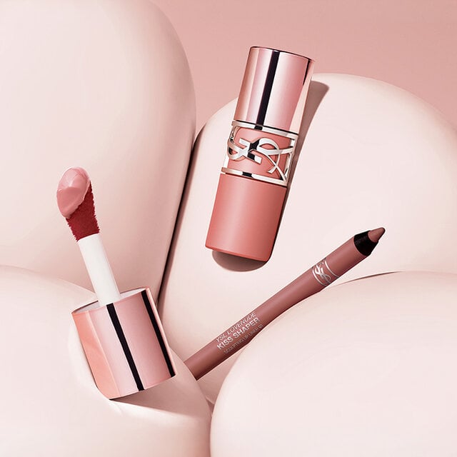 YSL LOVENUDE Kiss Shaper pencils and Lip Blusher lipsticks in various shades, arranged on a pink background.