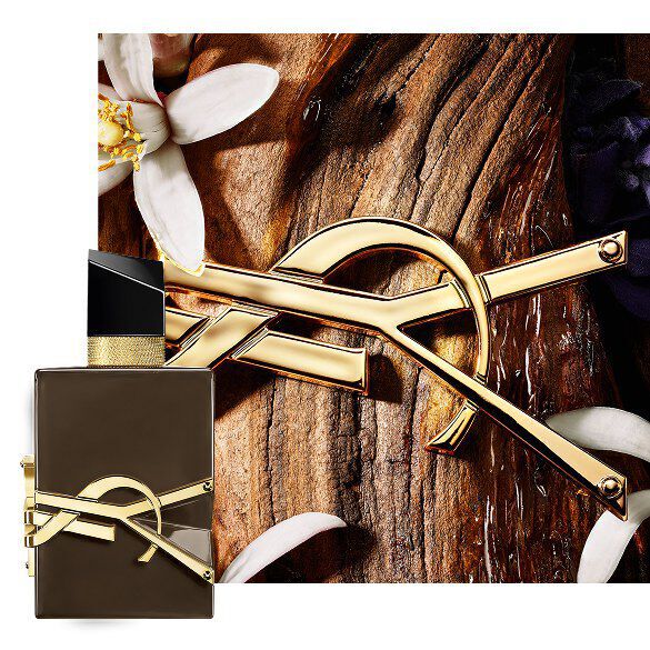 Libre Eau de Parfum bottle styled with gold chain and purple flowers on a dark background.