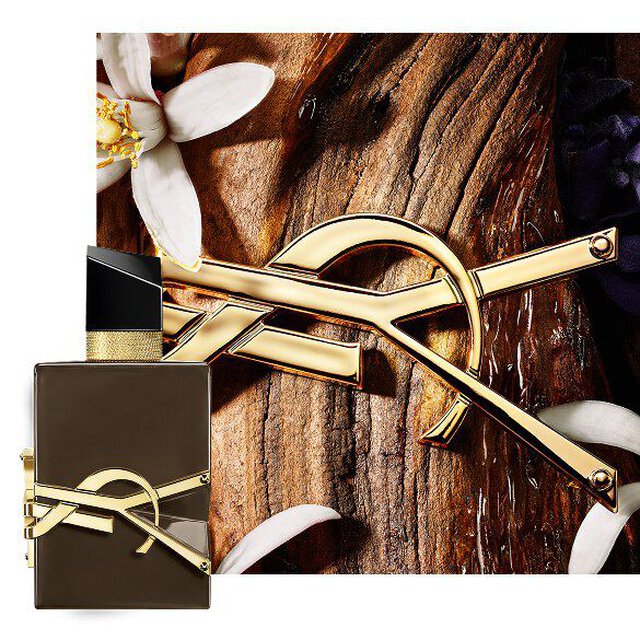 Libre Eau de Parfum bottle styled with gold chain and purple flowers on a dark background.