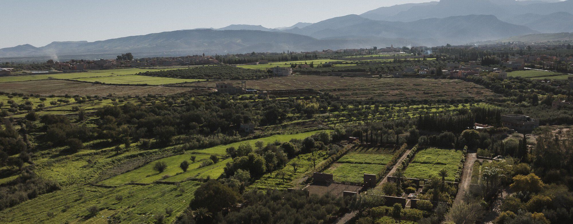 YSL For The Future: Aerial view of the Ourika Community Gardens,
lush green fields, trees, and mountains under a clear sky.