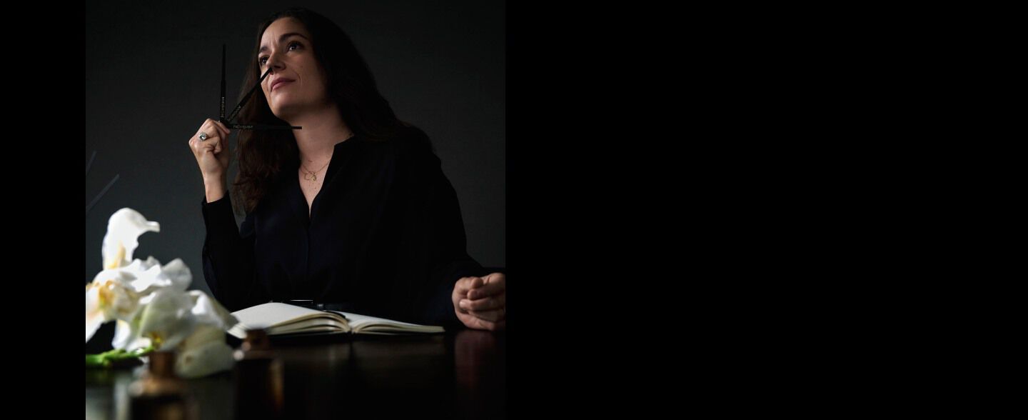 Perfumer Juliette Karagueuzoglou smelling fragrance while holding a blotter strip, seated at a table.